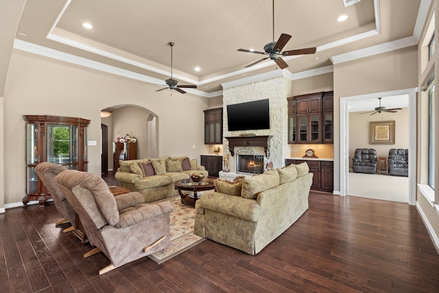 living area featuring a tray ceiling, arched walkways, a stone fireplace, and a ceiling fan