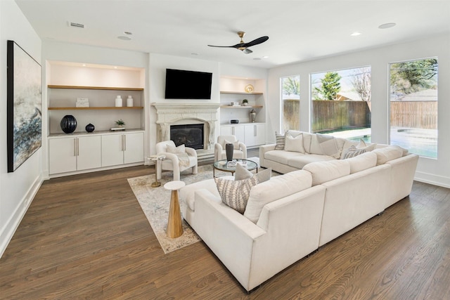 living area with dark wood finished floors, built in features, ceiling fan, and a glass covered fireplace