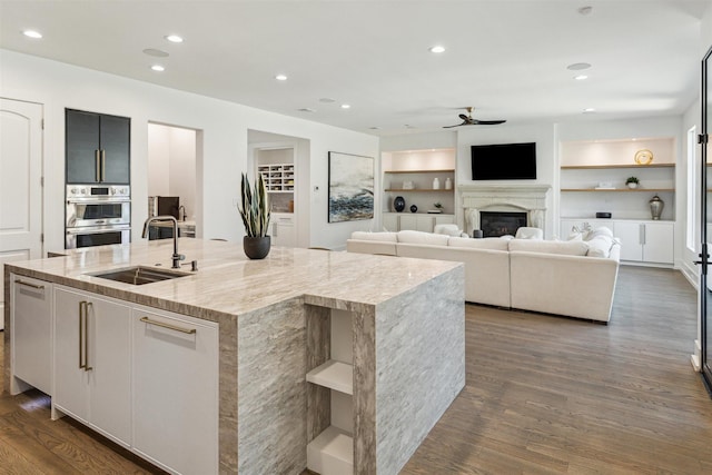 kitchen with an island with sink, a sink, dark wood-style floors, stainless steel double oven, and ceiling fan