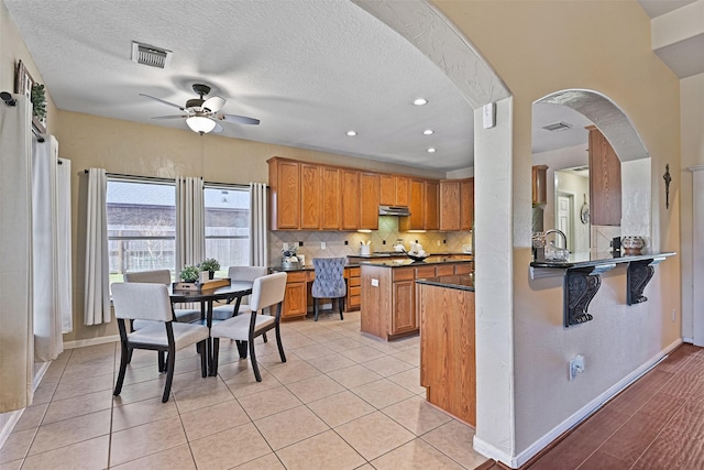 kitchen featuring visible vents, decorative backsplash, arched walkways, brown cabinetry, and a ceiling fan