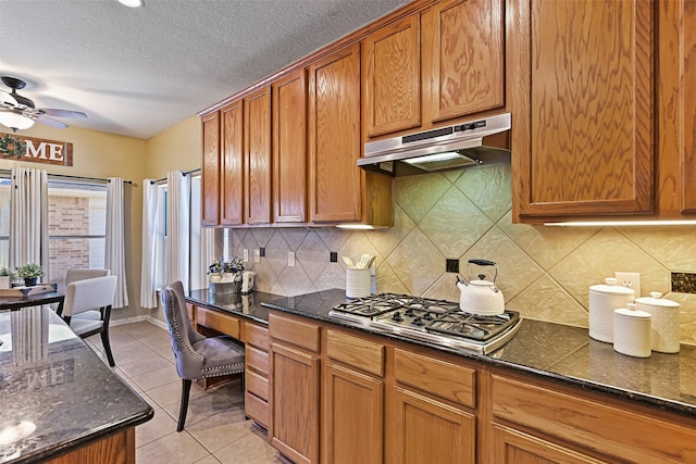 kitchen featuring under cabinet range hood, dark stone counters, stainless steel gas stovetop, light tile patterned floors, and ceiling fan