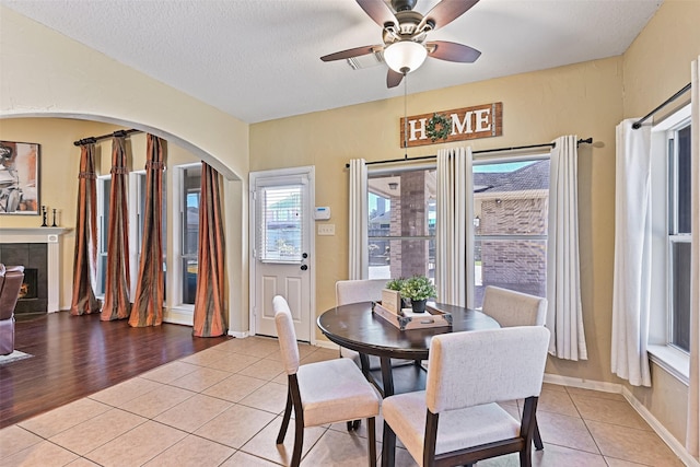 dining space with a tiled fireplace, light tile patterned floors, arched walkways, a textured ceiling, and a ceiling fan