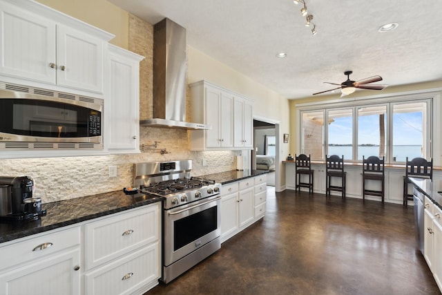 kitchen featuring ceiling fan, appliances with stainless steel finishes, white cabinetry, wall chimney range hood, and backsplash