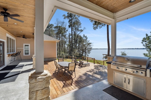 wooden terrace featuring a water view, an outdoor kitchen, a grill, outdoor dining area, and ceiling fan