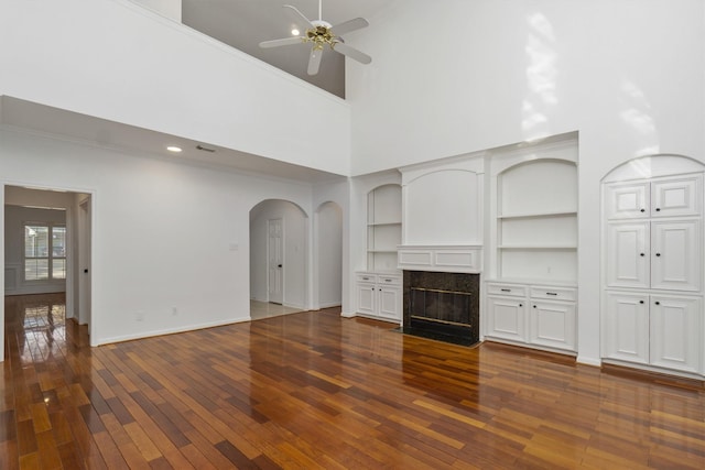 unfurnished living room featuring dark wood-type flooring, baseboards, ceiling fan, a fireplace, and arched walkways