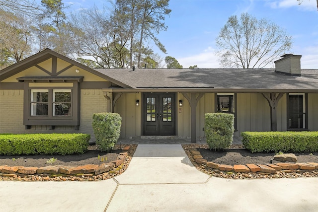 view of exterior entry with french doors, brick siding, roof with shingles, and a chimney