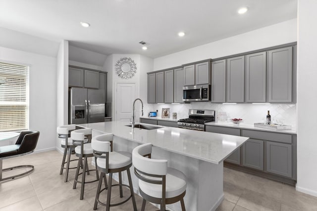 kitchen featuring a kitchen island with sink, gray cabinets, a sink, tasteful backsplash, and appliances with stainless steel finishes