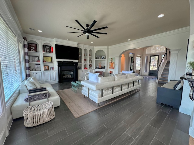 living area with crown molding, stairway, arched walkways, a glass covered fireplace, and a ceiling fan