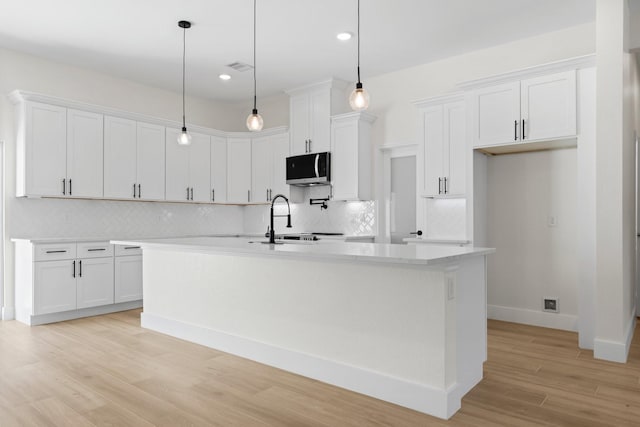 kitchen featuring stainless steel microwave, a center island with sink, light countertops, light wood-style floors, and white cabinetry