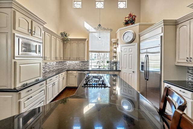 kitchen featuring tasteful backsplash, built in appliances, cream cabinets, a towering ceiling, and a sink