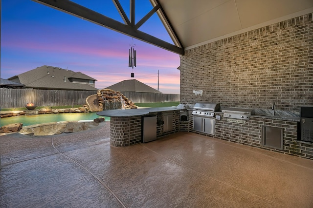 patio terrace at dusk with exterior kitchen, a fenced backyard, and a grill