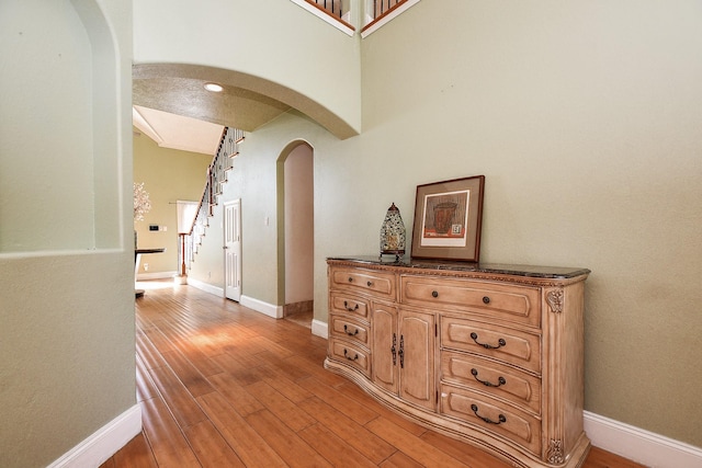 hall featuring baseboards, stairs, light wood-style flooring, a high ceiling, and arched walkways