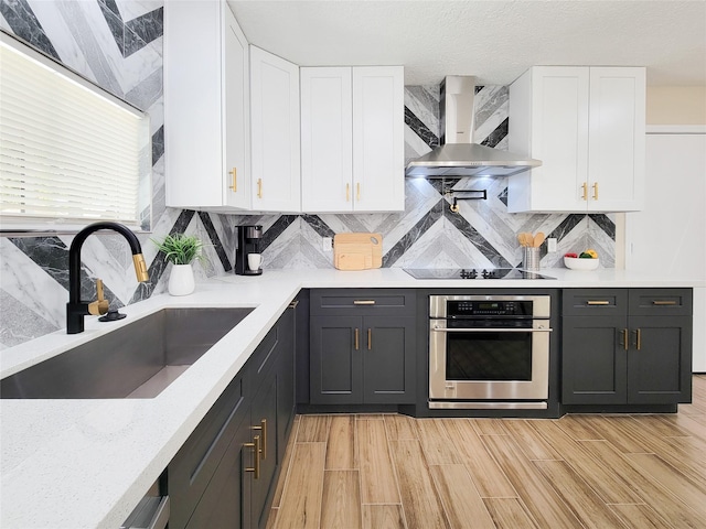 kitchen with a sink, white cabinets, stainless steel oven, wall chimney range hood, and black electric cooktop