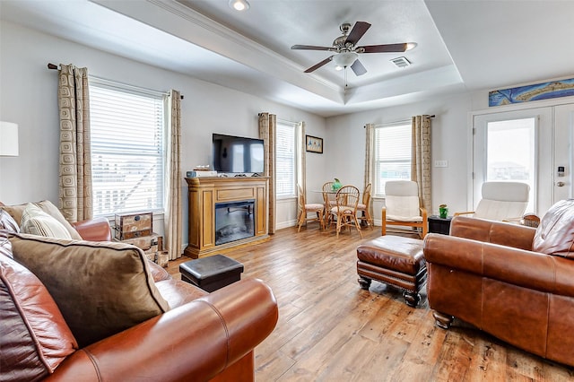 living area featuring visible vents, a tray ceiling, light wood-style floors, a glass covered fireplace, and a ceiling fan