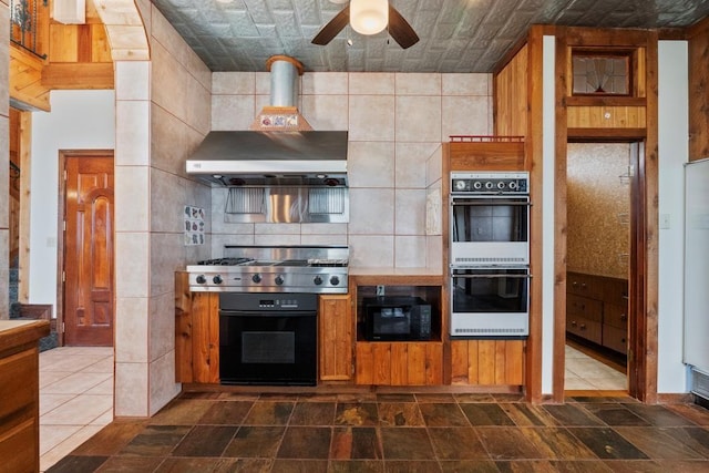 kitchen with multiple ovens, black microwave, stainless steel gas cooktop, wall chimney exhaust hood, and a ceiling fan