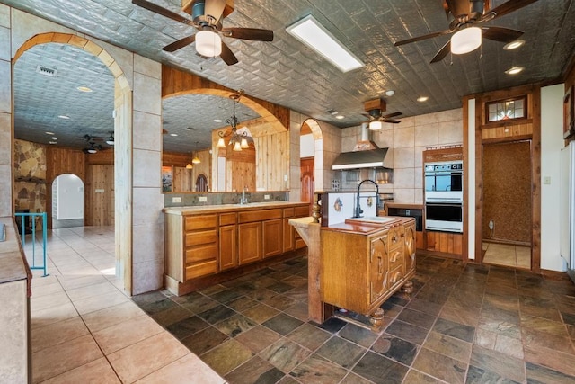 kitchen with wall chimney exhaust hood, double oven, and ceiling fan