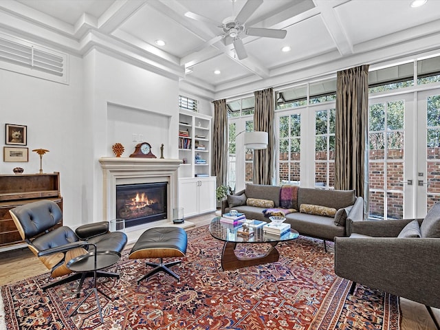 living area featuring a glass covered fireplace, built in features, coffered ceiling, and ceiling fan