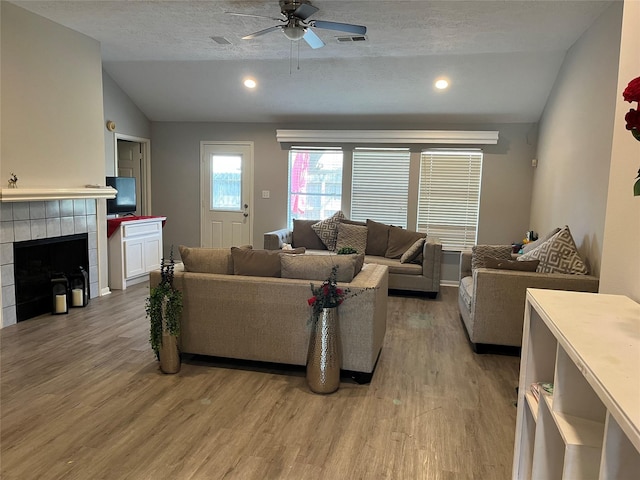 living room with ceiling fan, a tiled fireplace, light wood-type flooring, lofted ceiling, and a textured ceiling