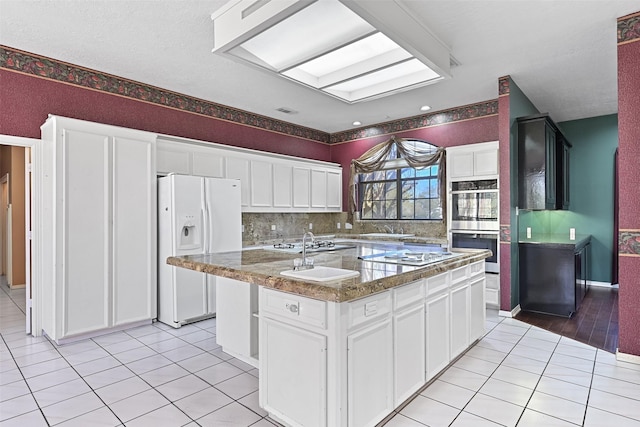 kitchen with tasteful backsplash, light tile patterned floors, white cabinets, white appliances, and a kitchen island with sink