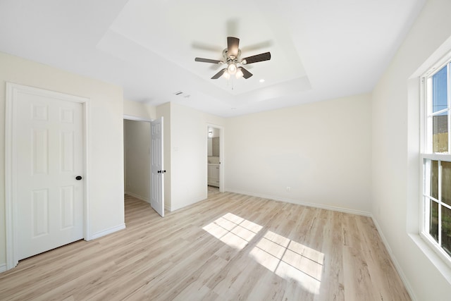 unfurnished bedroom featuring baseboards, ensuite bath, a tray ceiling, ceiling fan, and light wood-type flooring