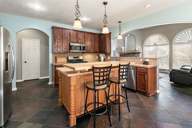 kitchen featuring stone tile floors, a peninsula, butcher block countertops, decorative backsplash, and appliances with stainless steel finishes