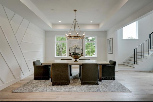 dining space featuring a tray ceiling, stairway, light wood-style floors, a decorative wall, and a chandelier