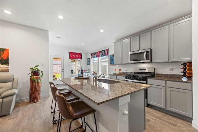 kitchen featuring gray cabinetry, a center island with sink, a sink, appliances with stainless steel finishes, and stone counters