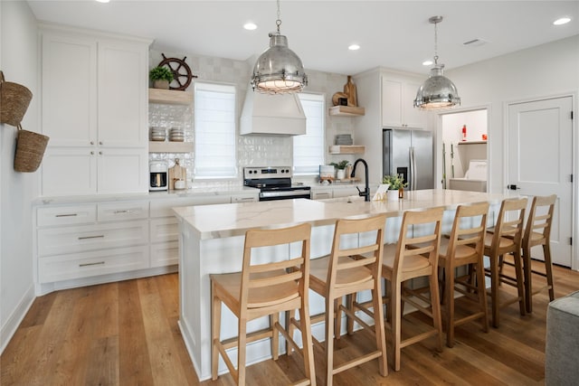 kitchen featuring open shelves, custom range hood, appliances with stainless steel finishes, light wood-style floors, and a kitchen island with sink