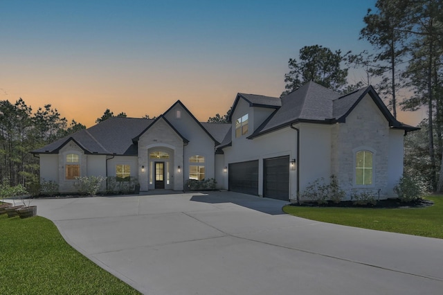 french country style house with a front yard, an attached garage, stucco siding, concrete driveway, and stone siding