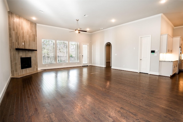 unfurnished living room featuring dark wood-type flooring, ornamental molding, a ceiling fan, a tiled fireplace, and arched walkways