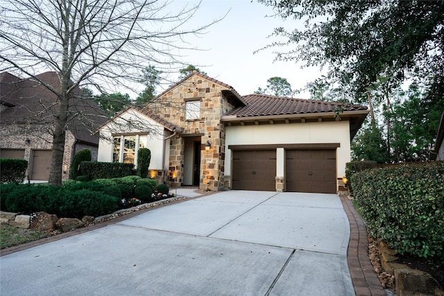 mediterranean / spanish house featuring an attached garage, a tiled roof, stucco siding, stone siding, and driveway