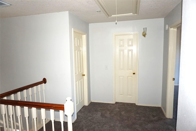 hallway featuring visible vents, an upstairs landing, a textured ceiling, dark colored carpet, and attic access