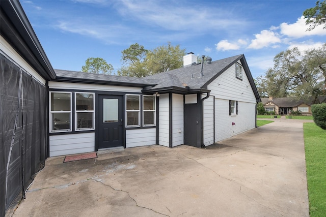 rear view of house featuring a patio area, a chimney, brick siding, and roof with shingles