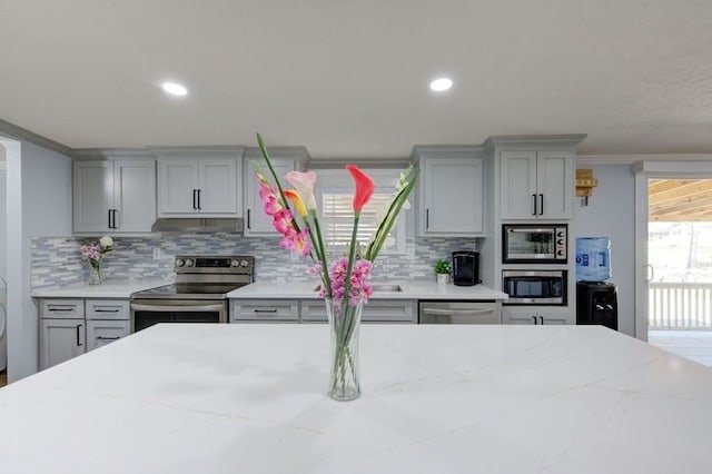 kitchen featuring backsplash, ornamental molding, light stone counters, gray cabinets, and appliances with stainless steel finishes