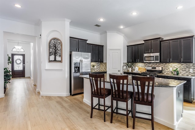 kitchen with a breakfast bar area, light wood-type flooring, dark stone counters, stainless steel appliances, and a kitchen island with sink