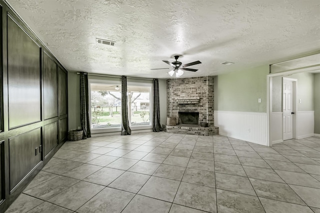 unfurnished living room with visible vents, a ceiling fan, wainscoting, a fireplace, and light tile patterned floors