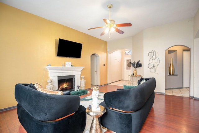 living area with baseboards, a lit fireplace, wood finished floors, arched walkways, and a ceiling fan