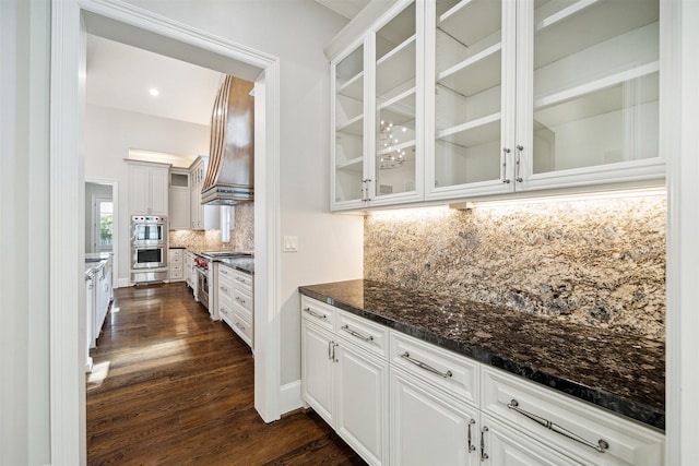 kitchen with backsplash, dark wood finished floors, appliances with stainless steel finishes, wall chimney range hood, and glass insert cabinets