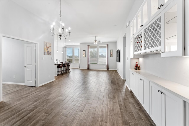 unfurnished living room featuring ceiling fan with notable chandelier, a dry bar, baseboards, and dark wood-style flooring