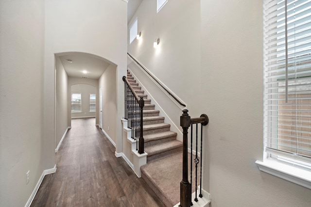 staircase featuring baseboards, arched walkways, wood finished floors, and a towering ceiling