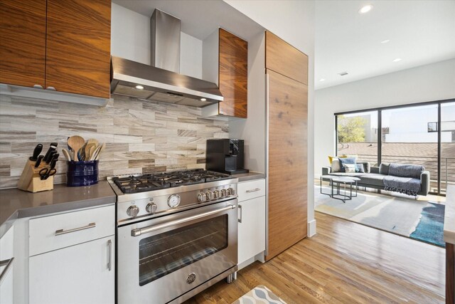 kitchen featuring light wood-style flooring, stainless steel stove, white cabinetry, wall chimney range hood, and backsplash