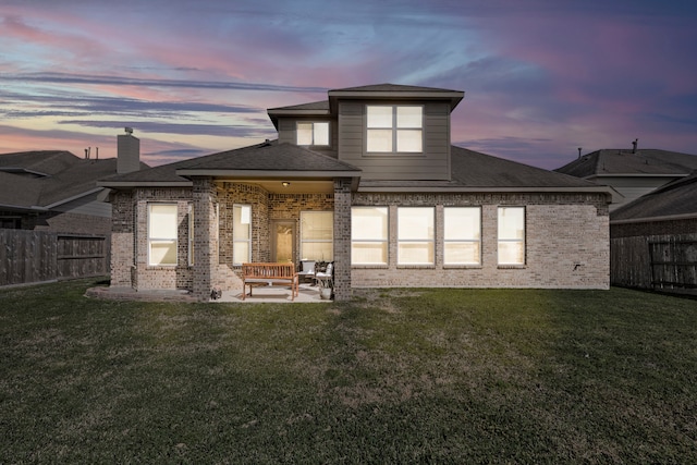 back of house at dusk with brick siding, fence, roof with shingles, a yard, and a patio area