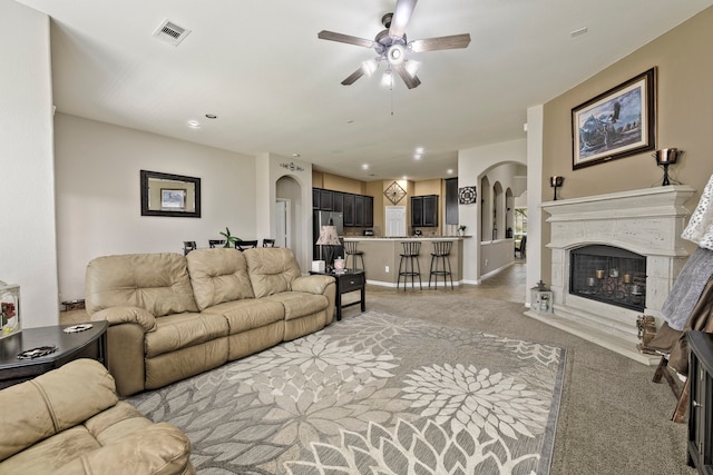 living area featuring a ceiling fan, visible vents, a fireplace with raised hearth, arched walkways, and light carpet