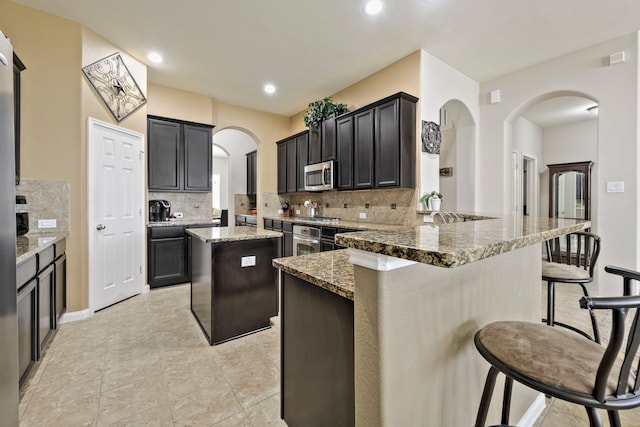 kitchen featuring a kitchen bar, light stone counters, stainless steel appliances, arched walkways, and a peninsula