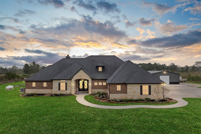 french country home featuring stone siding, roof with shingles, concrete driveway, a front yard, and a chimney