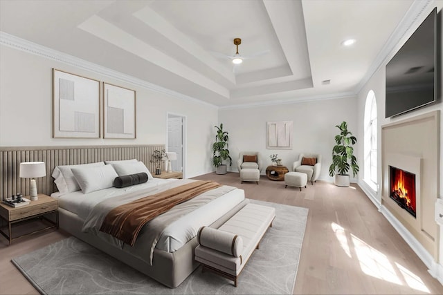 bedroom featuring light wood finished floors, ornamental molding, a warm lit fireplace, a raised ceiling, and a ceiling fan