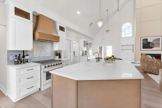 kitchen with custom exhaust hood, an island with sink, high end stove, glass insert cabinets, and open floor plan