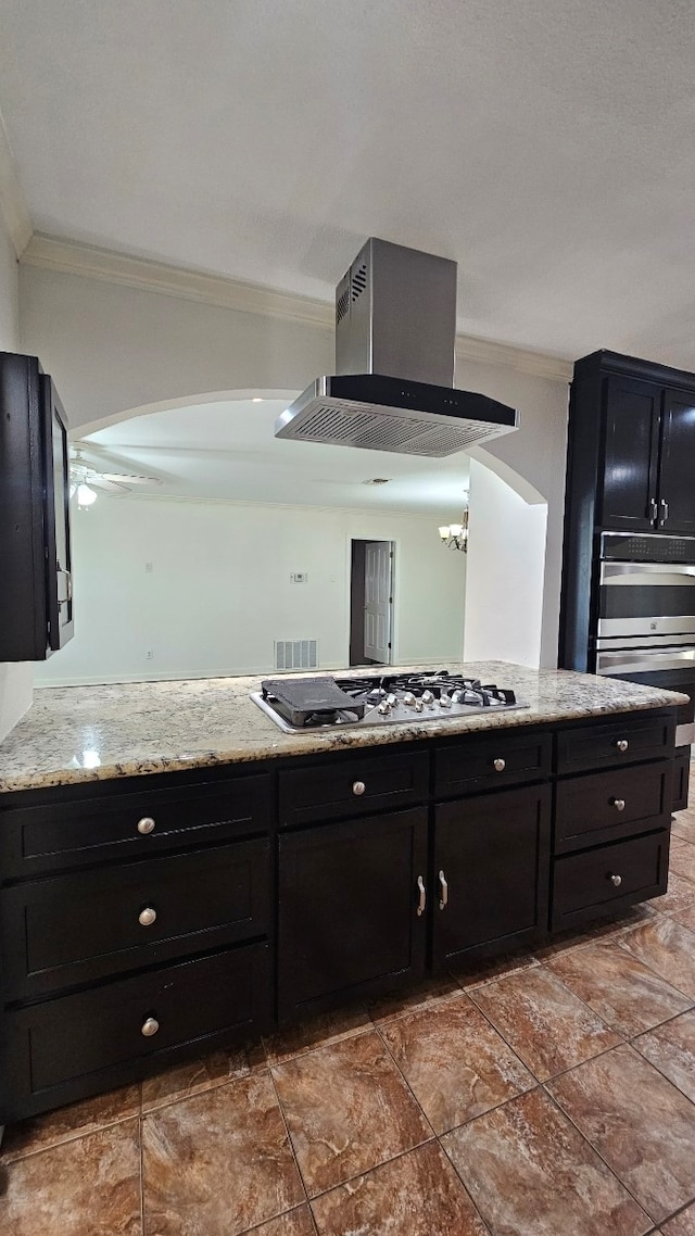 kitchen featuring crown molding, stainless steel gas cooktop, dark cabinetry, light stone counters, and wall chimney range hood