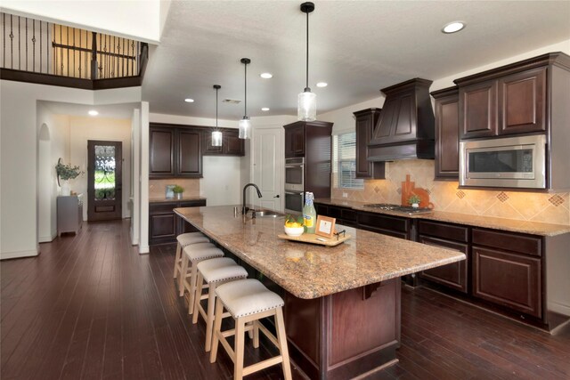 kitchen with dark brown cabinets, appliances with stainless steel finishes, light stone counters, a sink, and custom exhaust hood