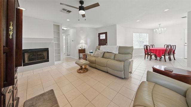 living room featuring visible vents, built in shelves, a tiled fireplace, light tile patterned floors, and ceiling fan with notable chandelier